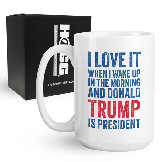 White mug with text 'I love it when I wake up in the morning and Donald Trump is president' in front of a black box with 'HOGGOUTFITTERS.COM' branding.