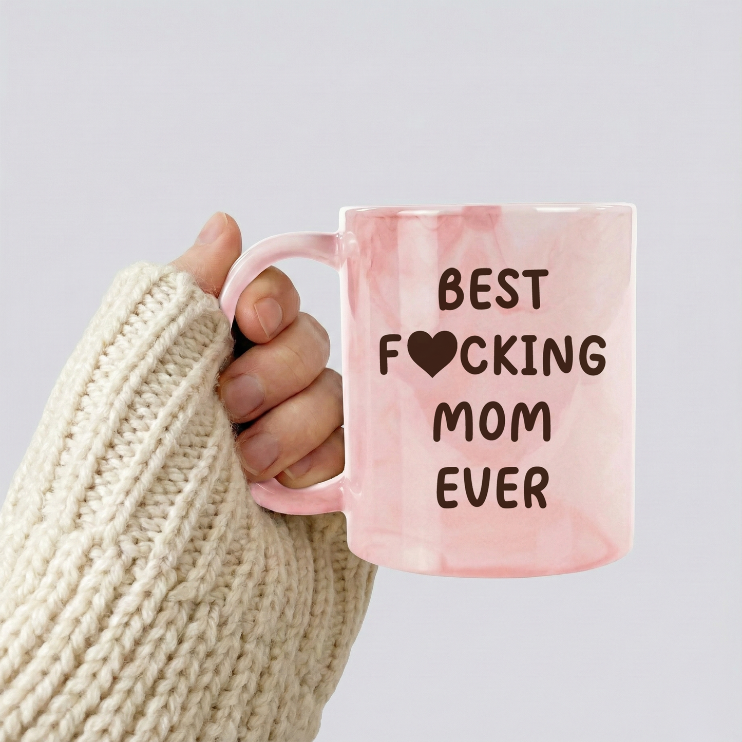 Hand holding a pink mug with 'BEST F*CKING MOM EVER' text against a light gray background
