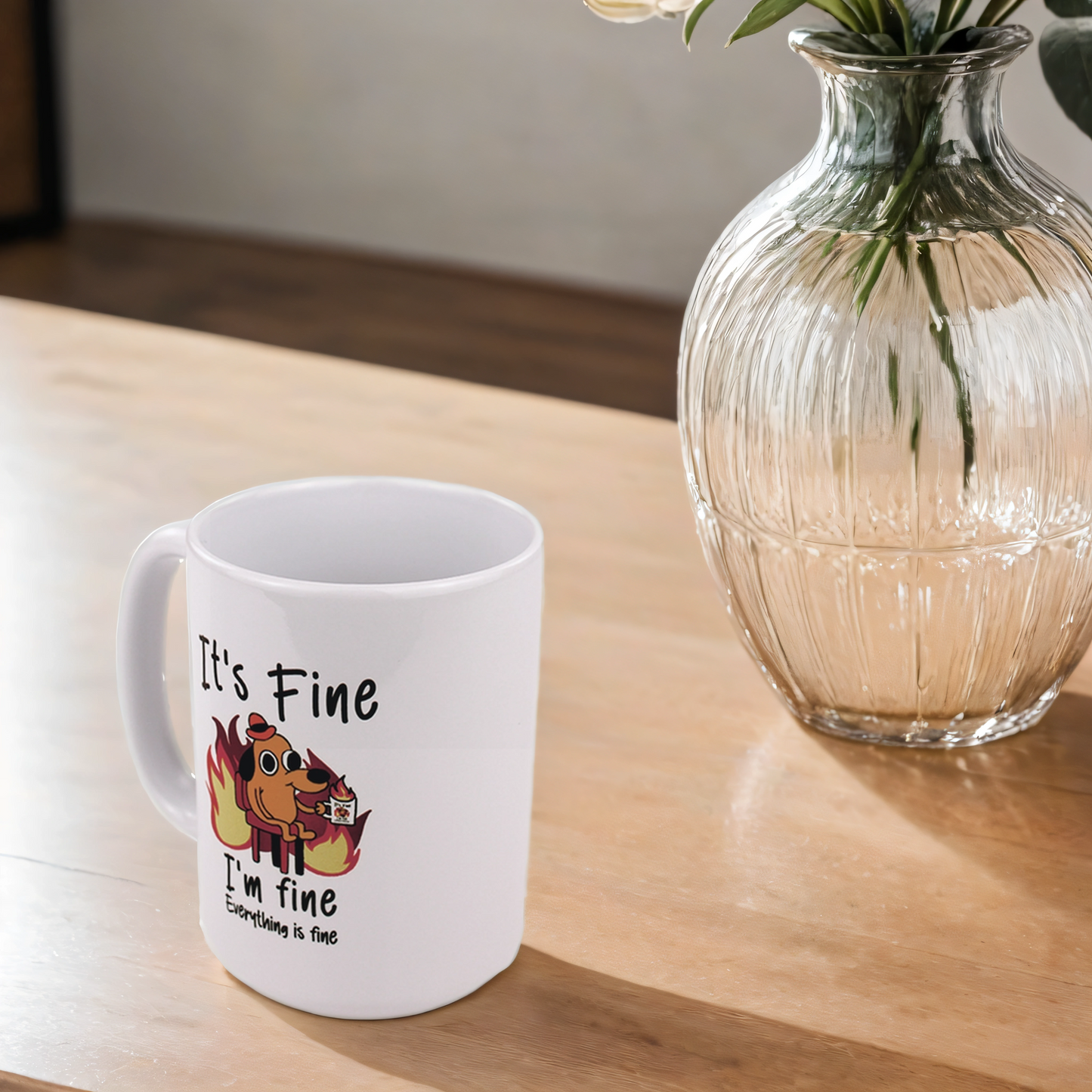 White mug with a cartoon design and text on a wooden table next to a glass vase with plants.